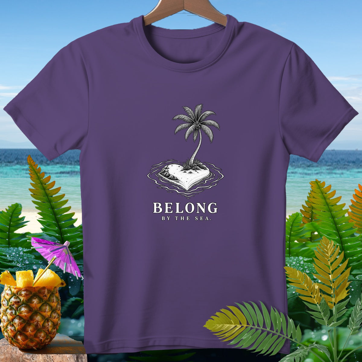 Purple t-shirt with heart-shaped island, palm tree, and text "BELONG BY THE SEA."