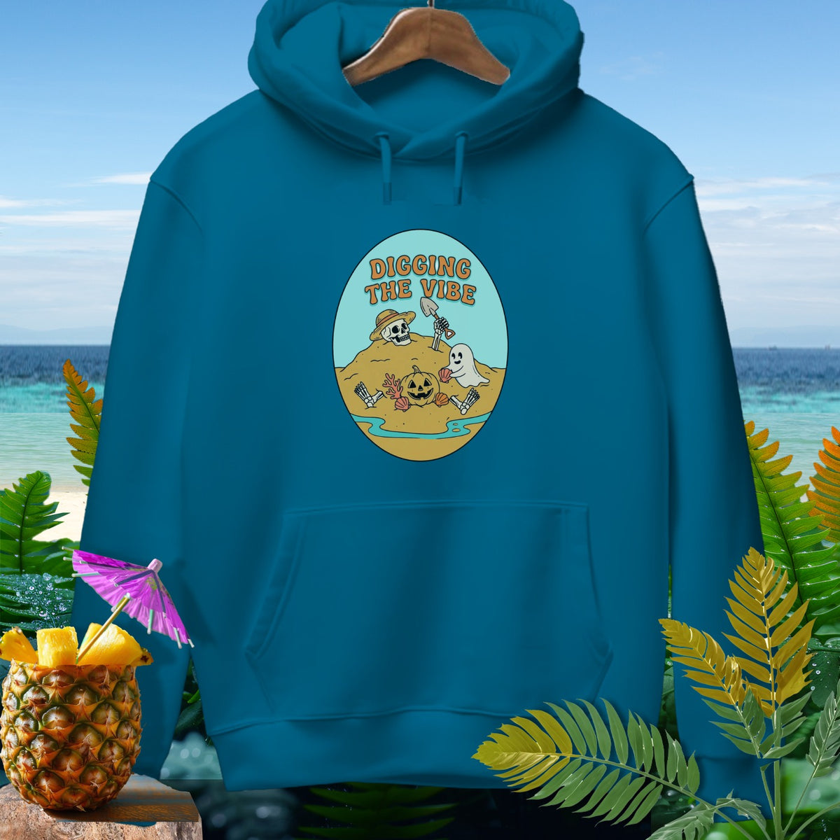 Teal hoodie with oval graphic depicting skeleton, ghost, and pumpkin digging on a beach.