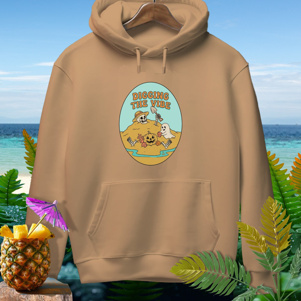 Tan hoodie with oval graphic of skeleton, ghost, and pumpkin digging on a beach.