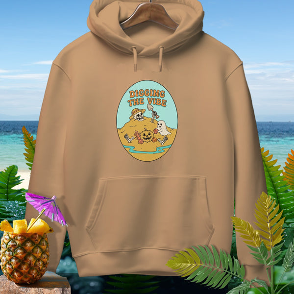 Tan hoodie with oval graphic of skeleton, ghost, and pumpkin digging on a beach.