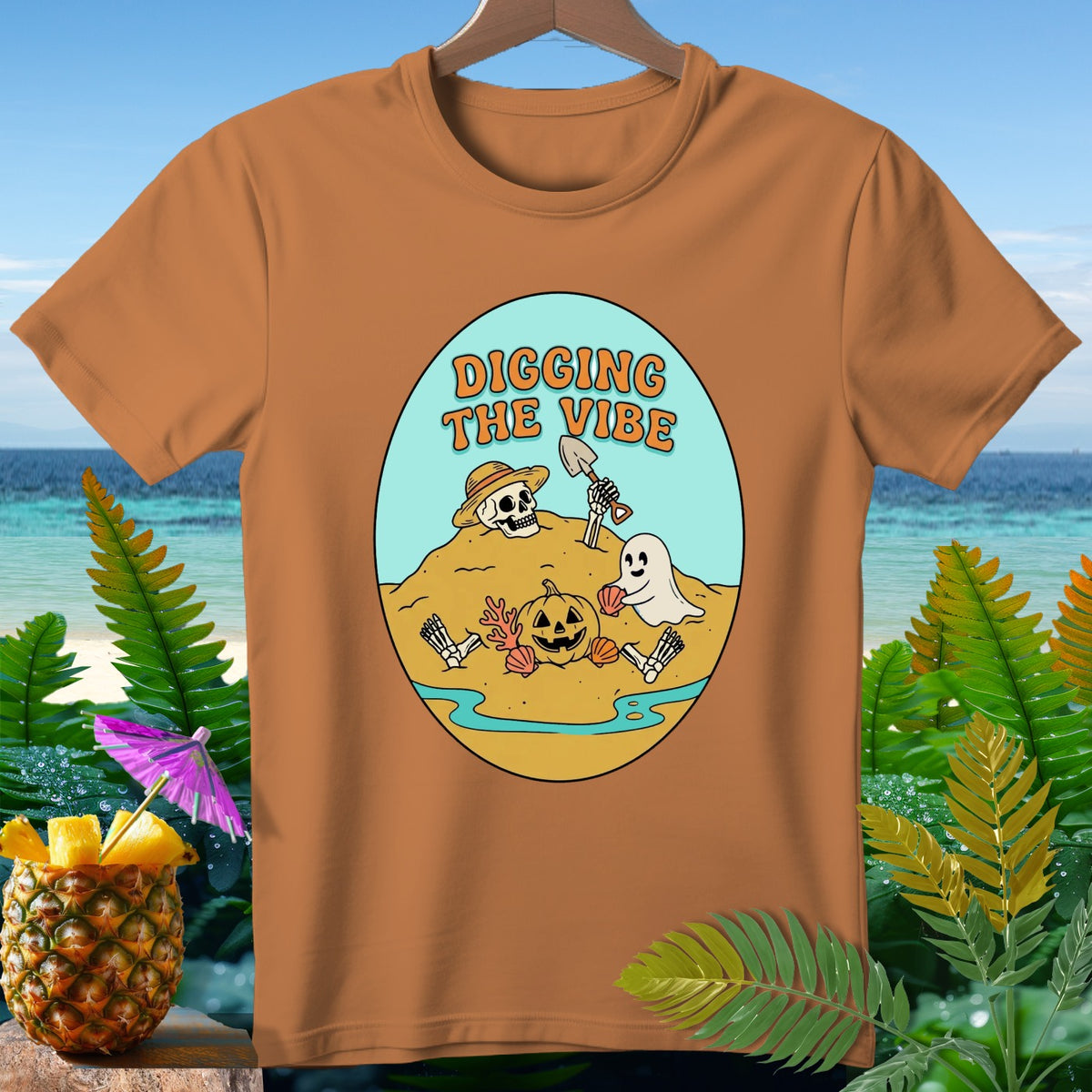 Brown t-shirt with cartoon illustration of skeleton digging on beach with ghost and pumpkin