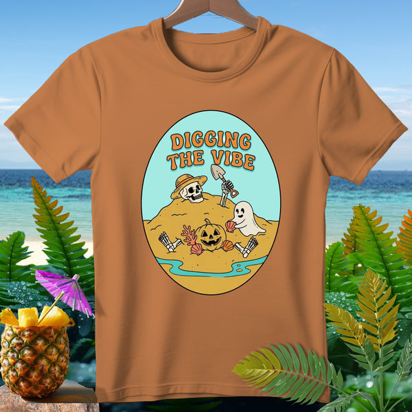 Brown t-shirt with cartoon illustration of skeleton digging on beach with ghost and pumpkin