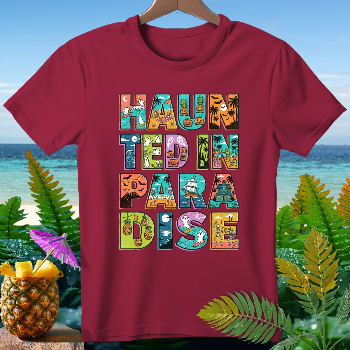 Maroon t-shirt with "HAUNTED IN PARADISE" text in colorful Halloween beach-themed letters.