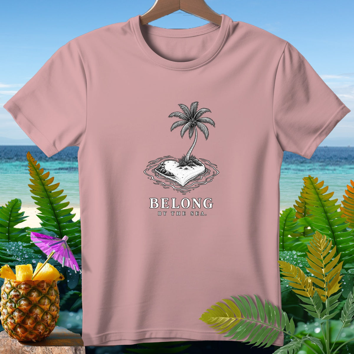 Pink t-shirt with a heart-shaped island, palm tree, and text "BELONG BY THE SEA."