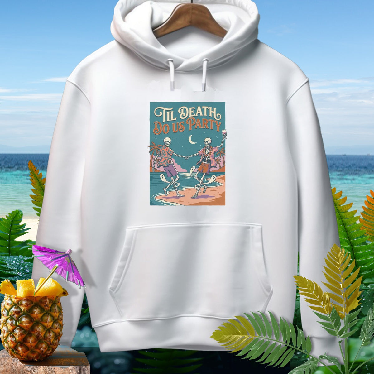 White hoodie with "Til Death Do Us Party" skeleton design on tropical beach.