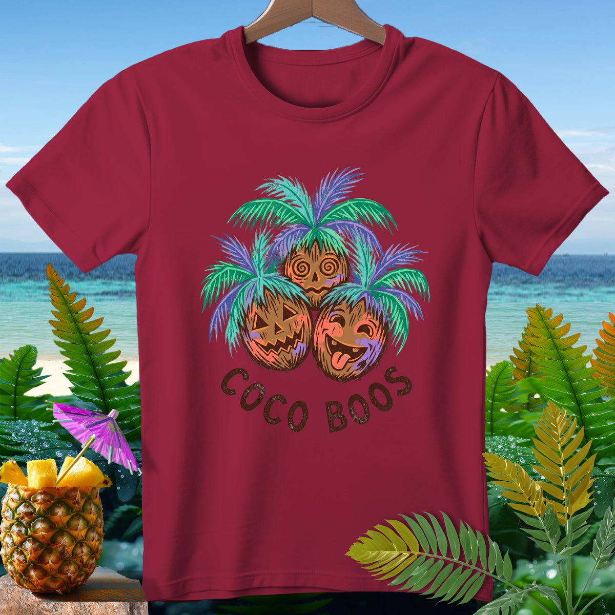 Burgundy t-shirt with three carved pumpkins under palm trees, text reads "Coco Boos".