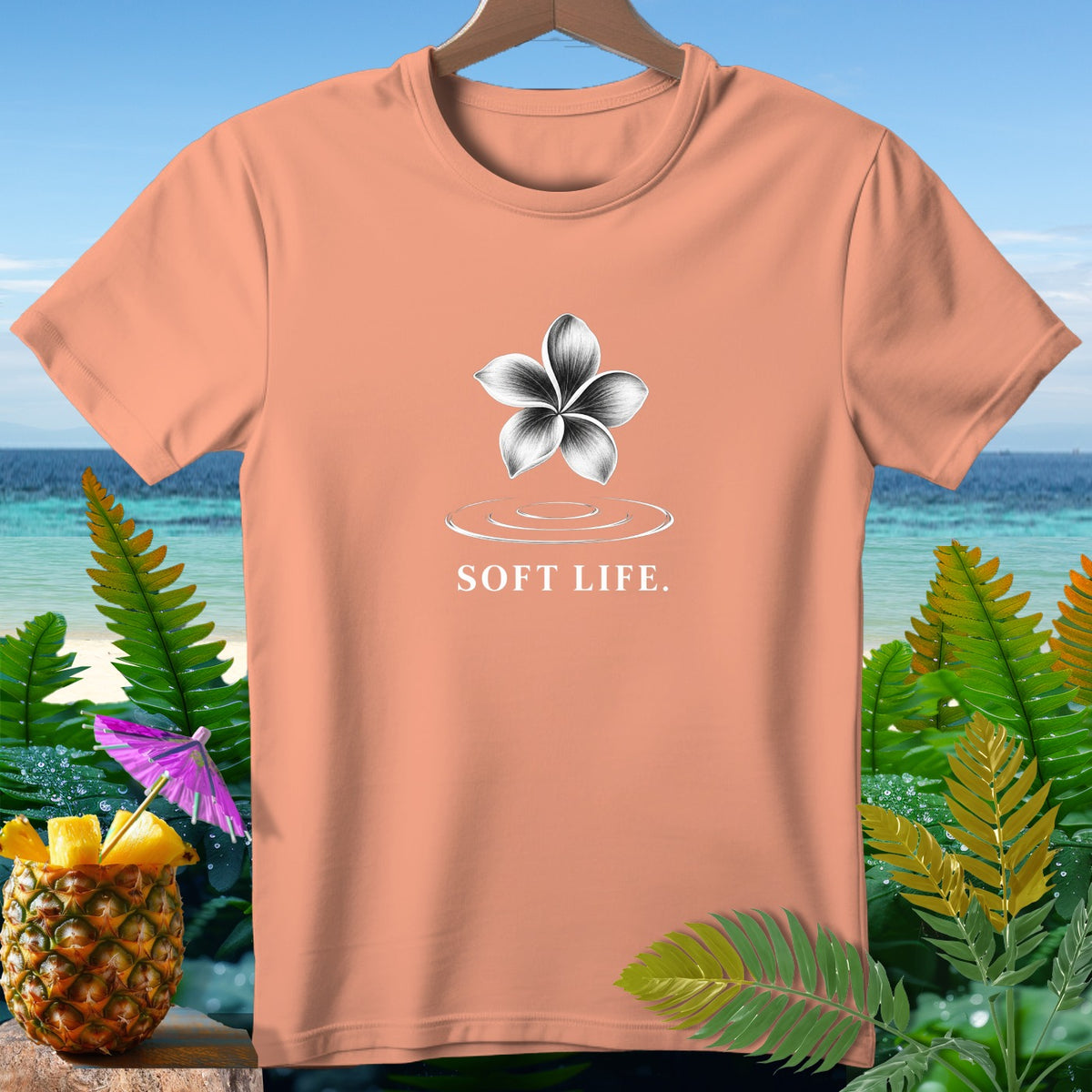 Peach t-shirt with flower graphic and "SOFT LIFE." text on beach background.