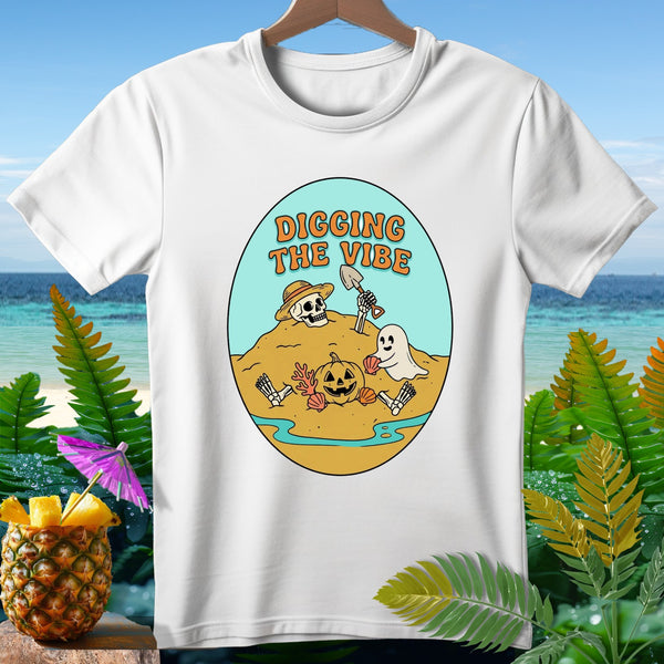 White t-shirt with a graphic of a skeleton, pumpkin, and ghost digging in the sand with text "Digging the Vibe."