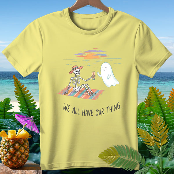 Yellow t-shirt with skeleton and ghost on a beach blanket, text reads "WE ALL HAVE OUR THING."