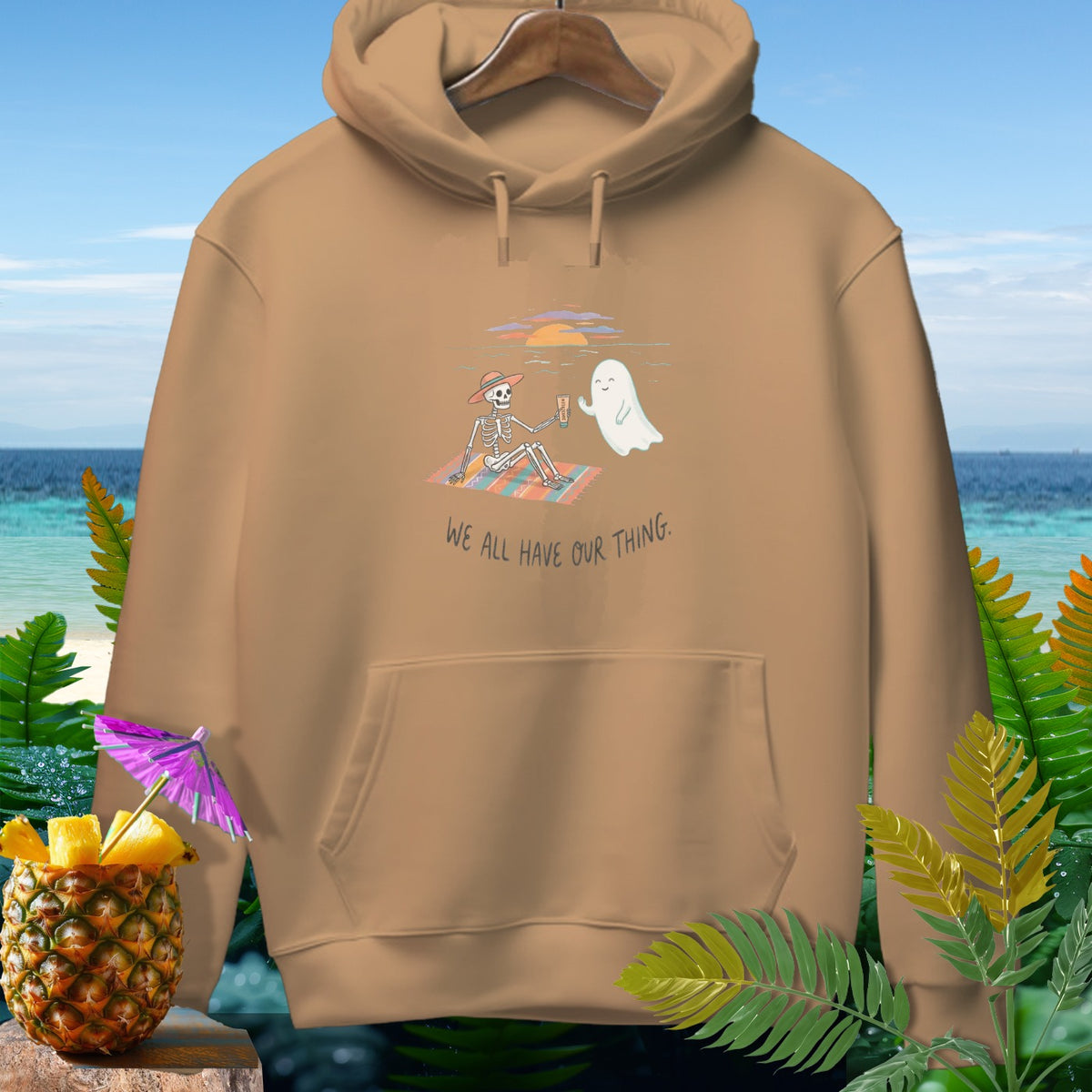 Tan hoodie with skeleton and ghost on a beach blanket, text "WE ALL HAVE OUR THING."