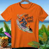 Orange t-shirt with witch, black cat, and broom surfing on rainbow waves with text 