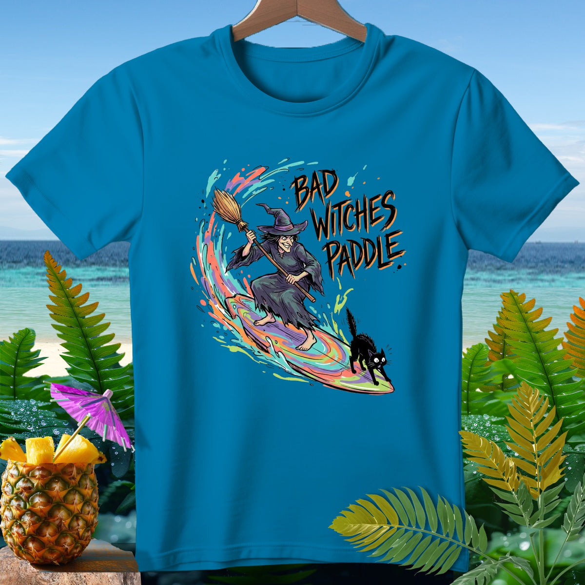 T-shirt graphic with witch and black cat surfing on a rainbow wave, text "Bad Witches Paddle".