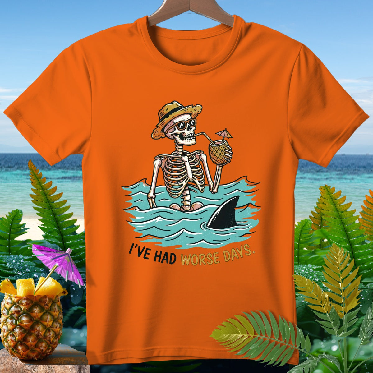 Orange t-shirt with a skeleton in a hat and sunglasses drinking from a pineapple, with a shark fin.