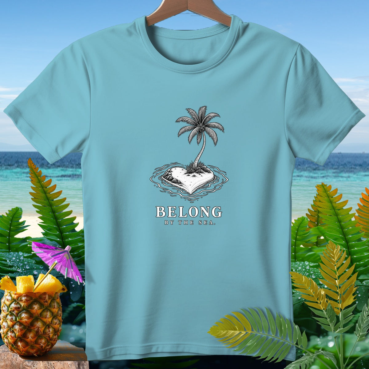 Light blue t-shirt with palm tree on heart-shaped island, text "BELONG BY THE SEA."