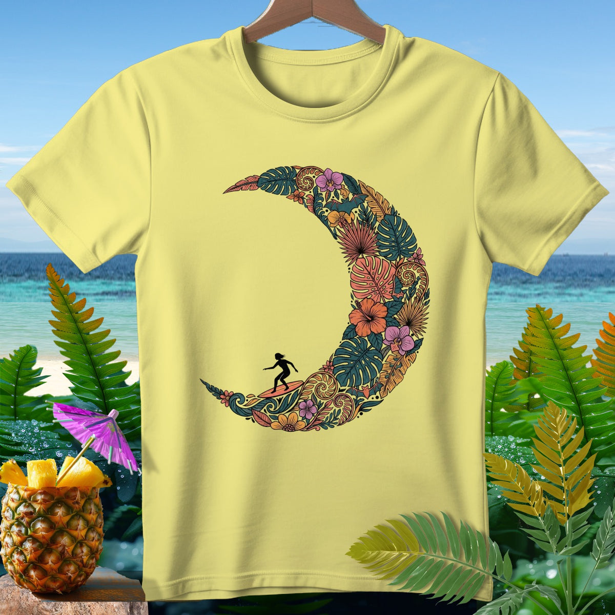 Yellow t-shirt with a crescent moon design filled with tropical leaves and flowers, featuring a surfer silhouette.