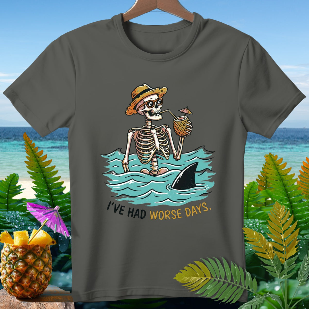 Skeleton with a straw in a pineapple drink in the ocean, shark fin nearby, text "I'VE HAD WORSE DAYS."
