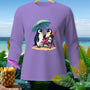 Purple long-sleeve shirt featuring two penguins on a beach under an umbrella.