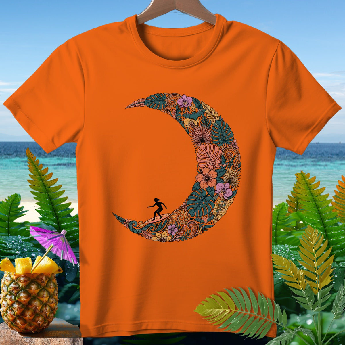 Orange t-shirt with a crescent moon graphic filled with tropical leaves and flowers, featuring a surfer.