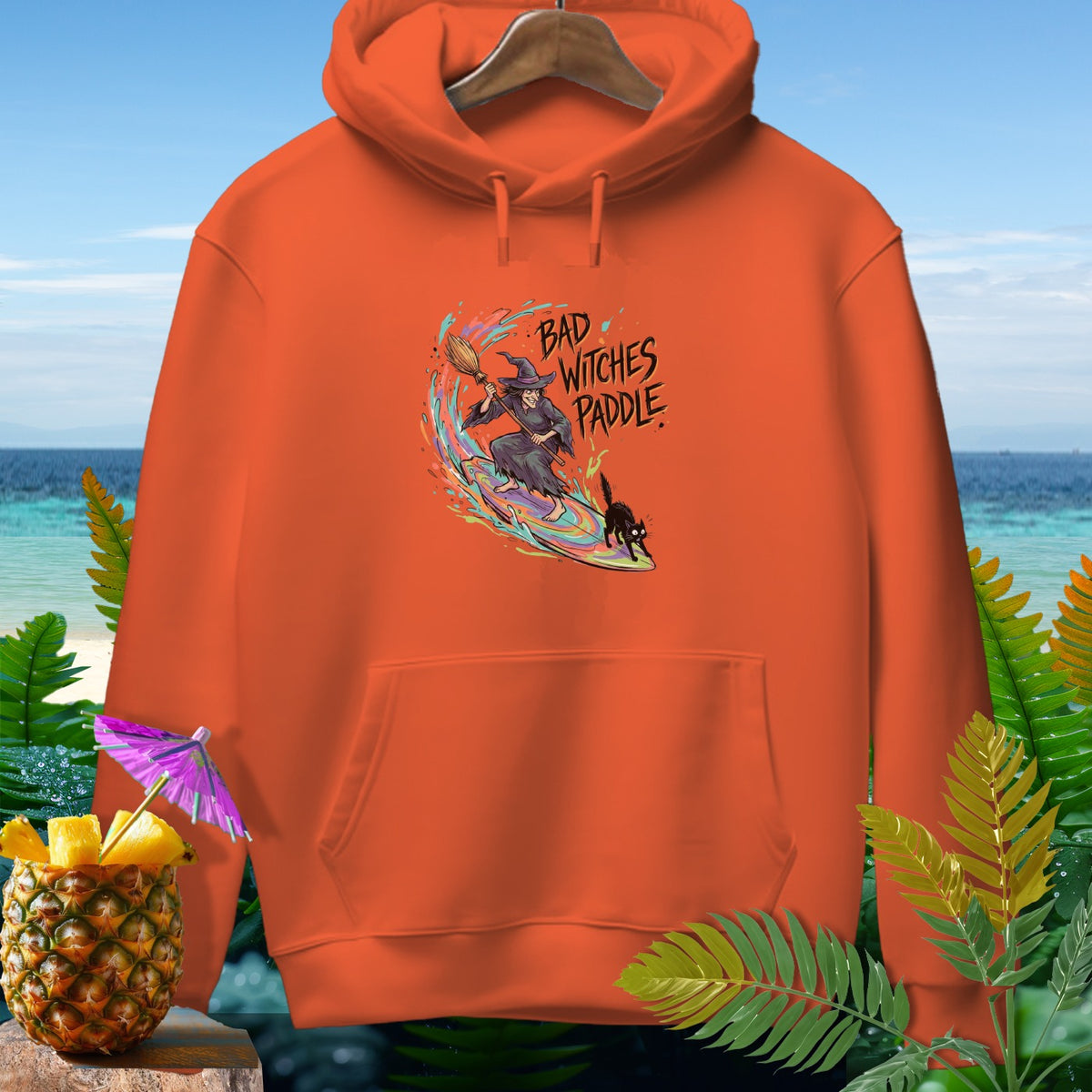 Orange hoodie with "Bad Witches Paddle" graphic of a witch and cat surfing.